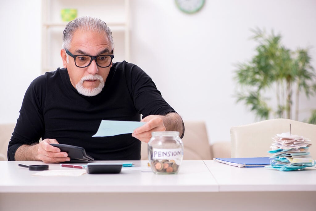 An older man wearing glasses looks at a document with a surprised expression while holding a wallet. A jar labeled 