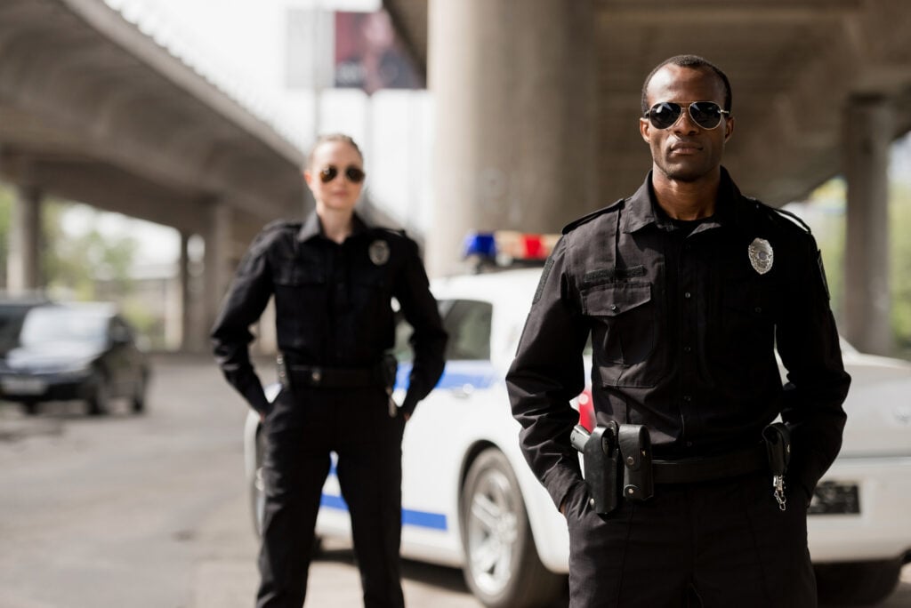 Two police officers in uniform stand in front of a police car under an overpass. Both officers are wearing sunglasses; the one in front has arms at his sides, and the one in back has hands in pockets. Their steadfast demeanor highlights that law enforcement is among those high paying jobs people don't want to do.