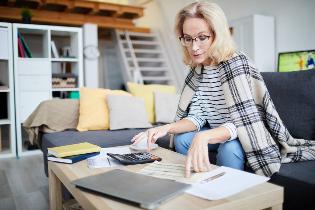 A woman with a blanket wrapped around her shoulders sits on a sofa, using a calculator and reviewing paperwork on a coffee table in the living room, concerned about things becoming too expensive.