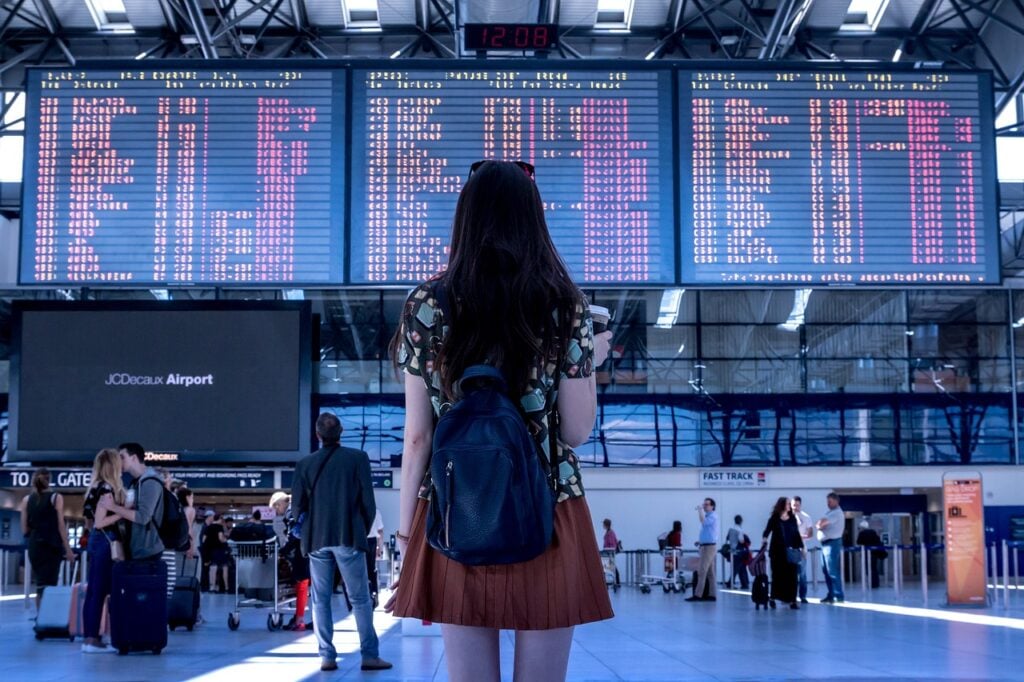 A woman with a backpack looks at a large airport departure board displaying various flight information. Other passengers and airport staff are visible in the background.