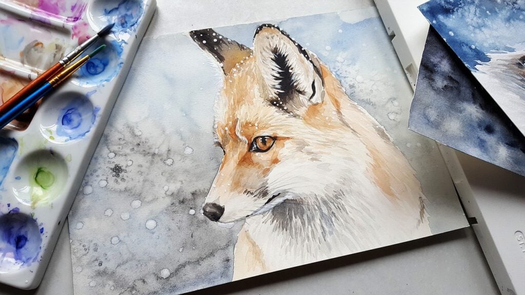 A watercolor painting of a fox is displayed alongside a watercolor palette and paintbrushes. The painting depicts the fox in a snowy setting with a detailed, expressive eye.