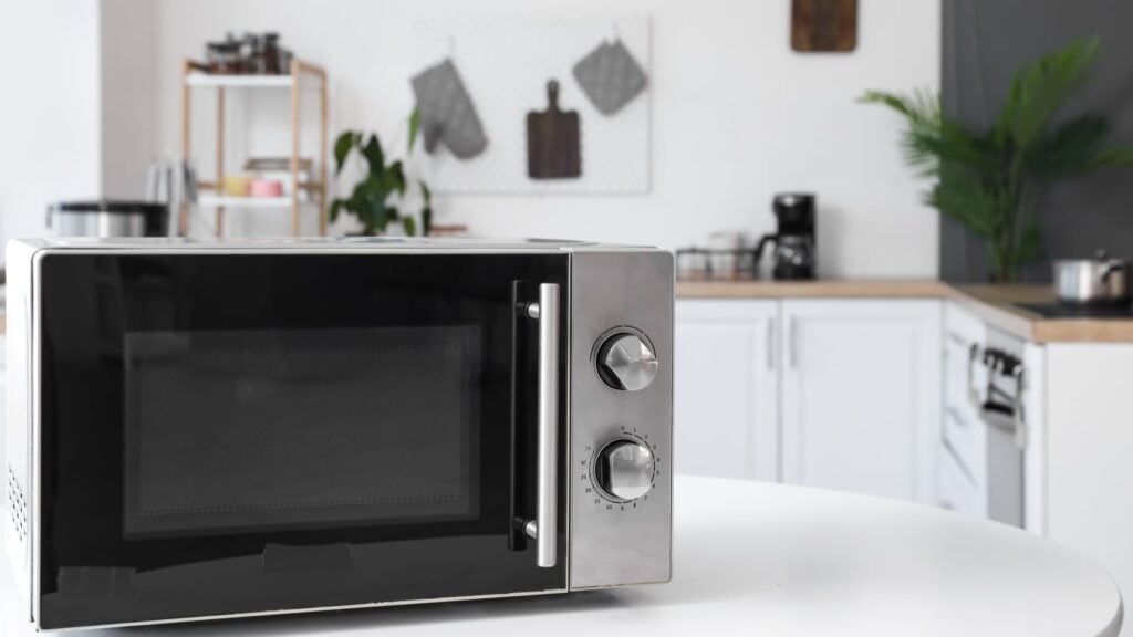 A microwave oven with a digital display and control dials sits on a white countertop in a modern kitchen with plants and various kitchen appliances in the background.