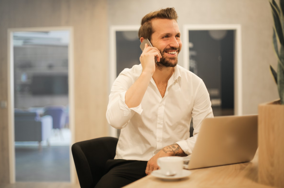 Man sitting in front of his laptop drinking a coffee on his phone smiling.