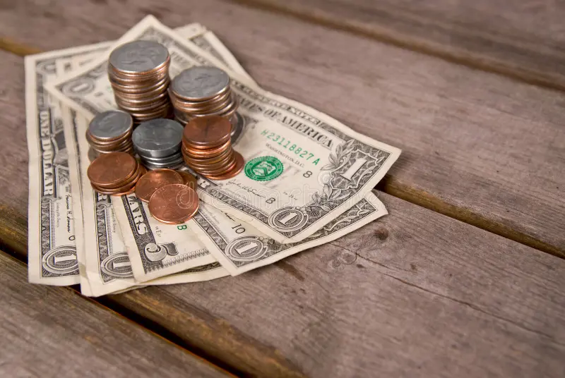 Stacks of coins and several US dollar bills on a wooden surface.