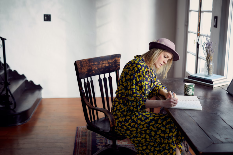 Woman wearing a hat writing in a notebook at her desk.