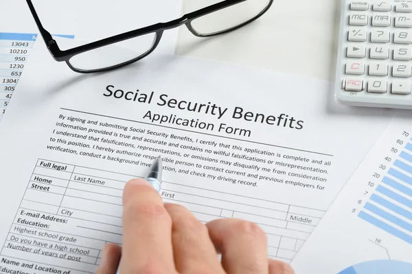 Close-up of a person filling out a Social Security Benefits Application Form on a desk with a pen, alongside financial documents, glasses, and a calculator.