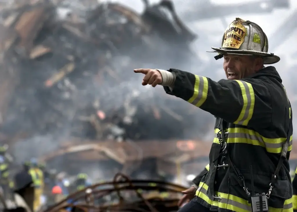 A firefighter in full gear points to the right amidst smoke and debris, with a large pile of rubble visible in the background.