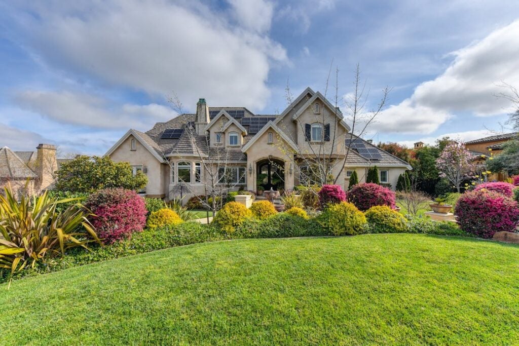 A large house with multiple gables and a landscaped front yard featuring a lush green lawn, flowering bushes, and small trees under a partly cloudy sky.