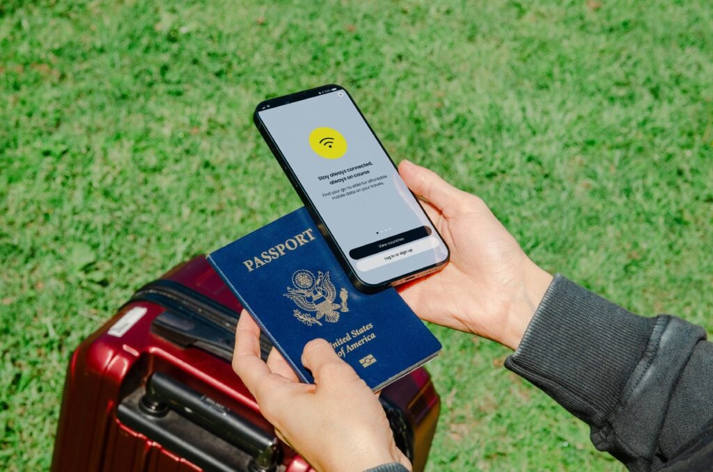 Person holding a smartphone showing a connectivity screen and a passport near a red suitcase on grass.