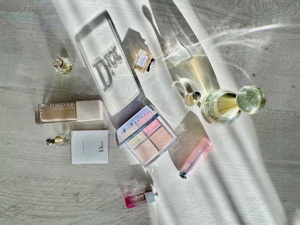 Various Dior makeup products and perfumes are laid out on a light-colored surface, including a highlighter palette, foundation, and lipsticks.