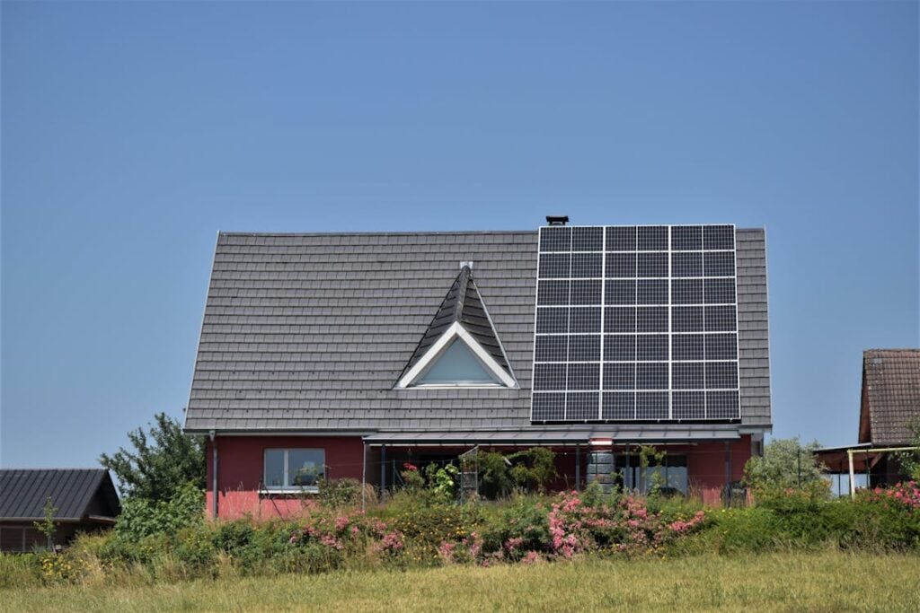 A house with a triangular window below its roof, fitted with solar panels on one side, surrounded by shrubs and plants, under a clear blue sky.