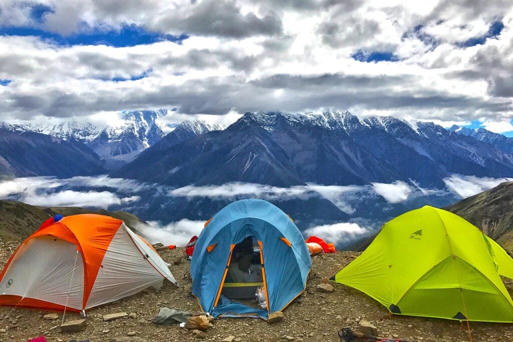 Three colorful tents set up on a rocky mountain slope with snow-capped peaks and a cloudy sky in the background.