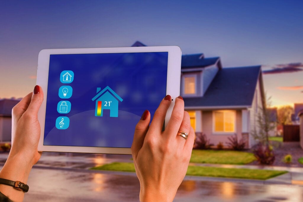 Person holding a tablet displaying a smart home control screen, with icons for home functions and a temperature of 21°C, in front of a modern house at dusk. Discover how to lower utility bills by optimizing your smart home settings effortlessly.