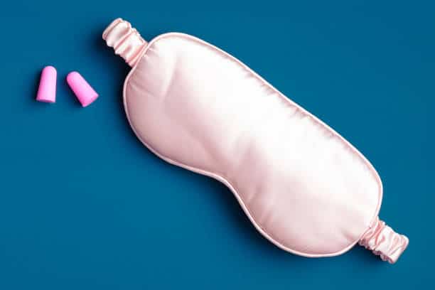 A pink sleep mask and a pair of pink earplugs are placed on a blue background.