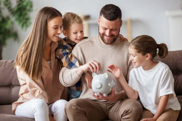 A family of four sitting on a couch, smiling and putting money into a gray piggy bank held by the father.