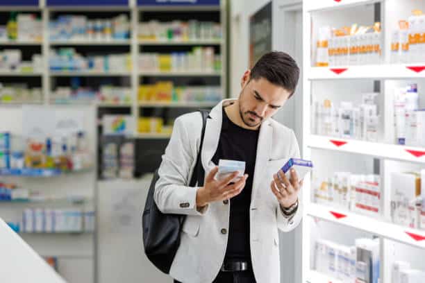 A man stands in a pharmacy holding and comparing two boxes of medication while looking closely at them.