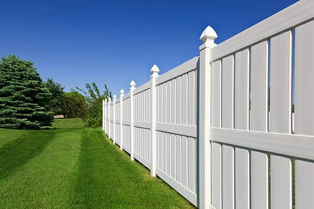 A long white vinyl fence runs along a green lawn with trees in the background under a clear blue sky.