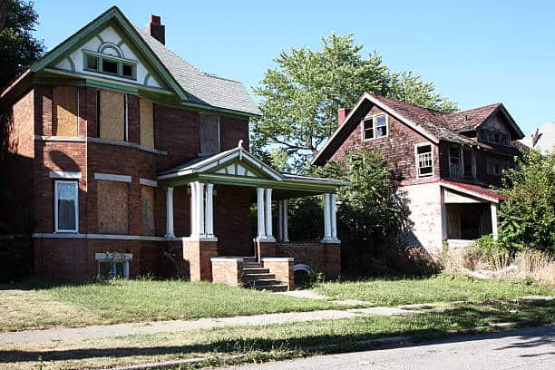 Two adjacent houses with distinct conditions: one well-maintained with a covered porch, the other dilapidated and overgrown with vegetation.