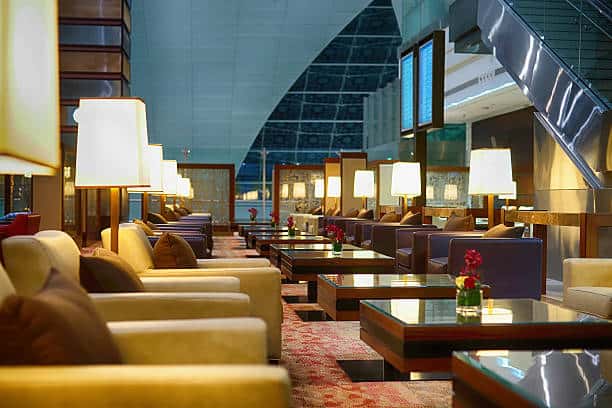 A modern airport lounge featuring rows of cushioned chairs and glass-top tables with lamps and flower arrangements. A staircase is visible on the right side. Large windows are in the background.