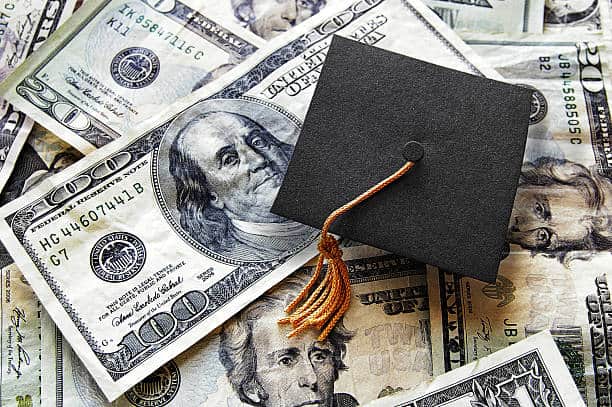 A graduation cap with a tassel sits on top of a pile of US dollar bills.