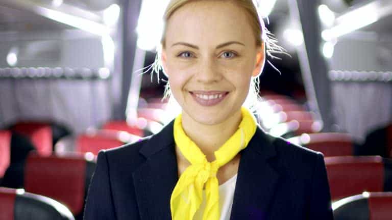 A person smiling, dressed in a professional uniform with a yellow scarf, standing in what appears to be the interior of a bus or an airplane cabin.