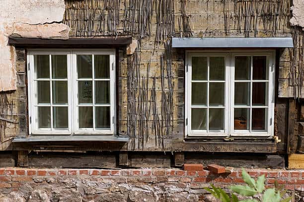 A weathered rustic wall with two white framed windows, partially covered with wooden slats and exposed bricks below.