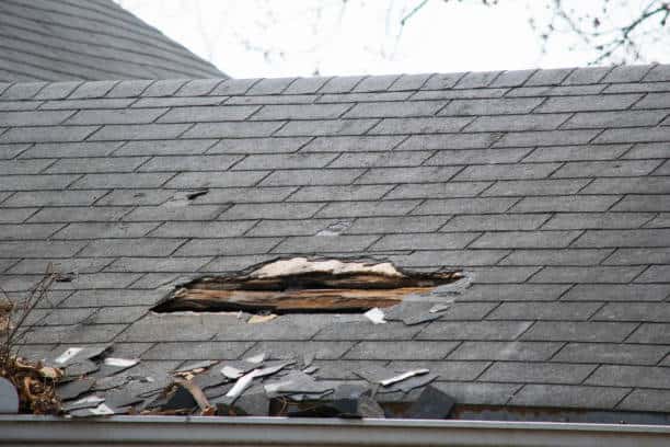 A damaged shingle roof with a large hole revealing the underlying structure. Debris and broken shingles are scattered around the damaged area.