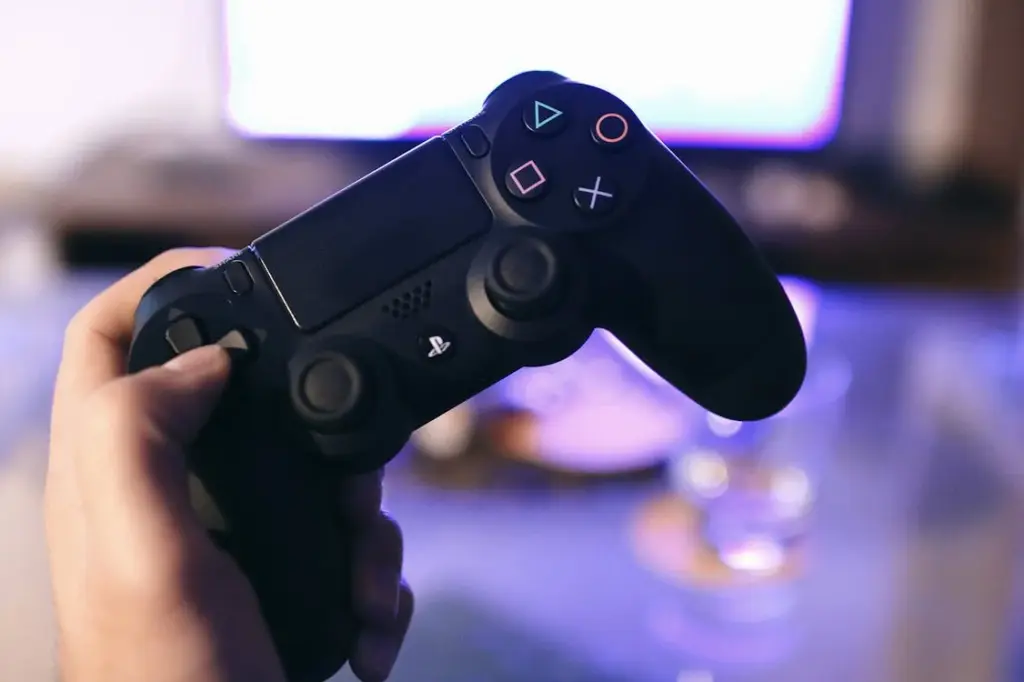 A hand holding a black PlayStation controller in front of a blurred TV screen.