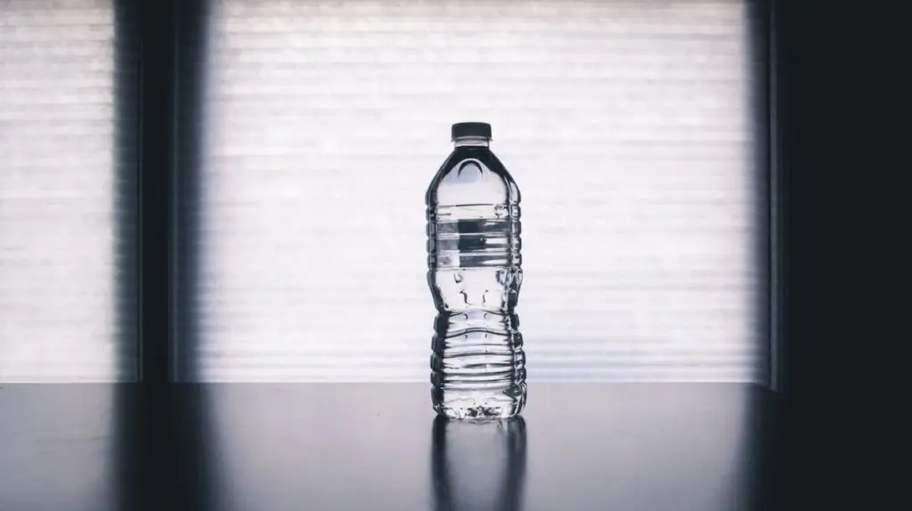 Clear plastic water bottle half-filled with water placed on a dark surface, with a blurred, bright background.