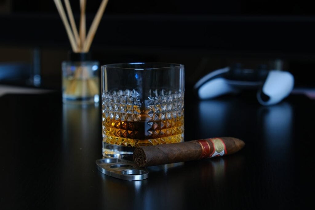 A glass of whiskey, a cigar, and a cigar cutter are placed on a dark wooden table. A diffuser and a white object are blurred in the background.