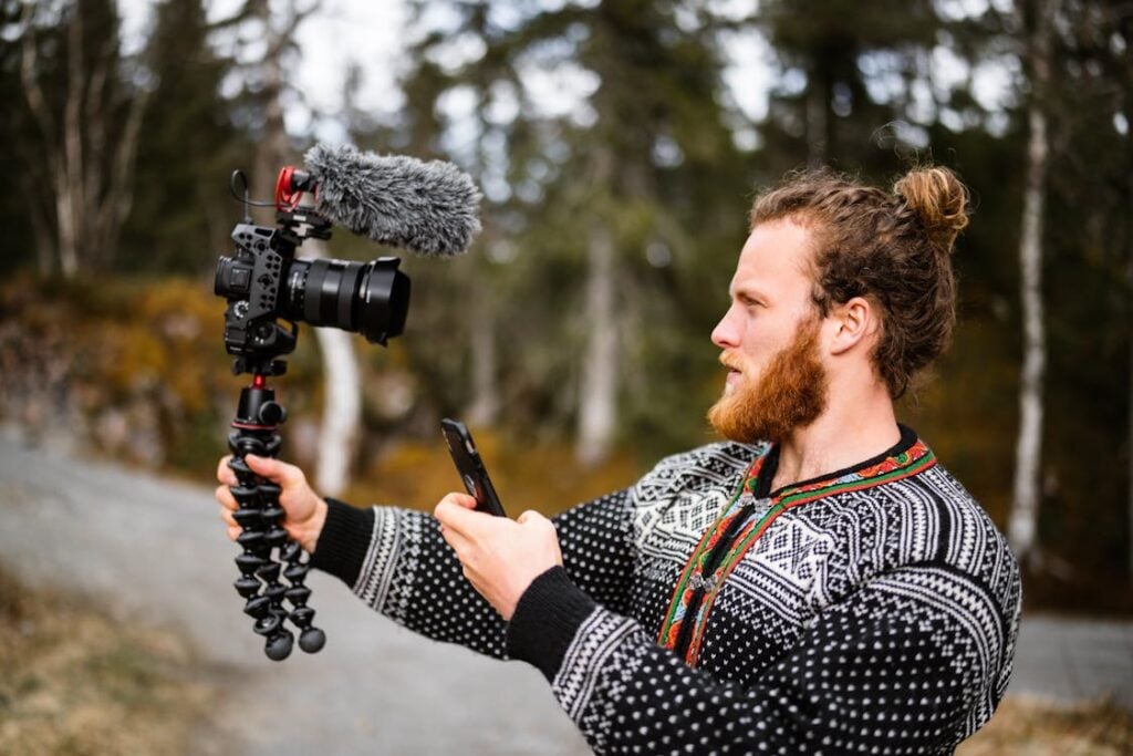 A person with a beard, wearing a patterned sweater, holds a camera with a microphone on a flexible tripod in one hand and a smartphone in the other, standing outdoors.