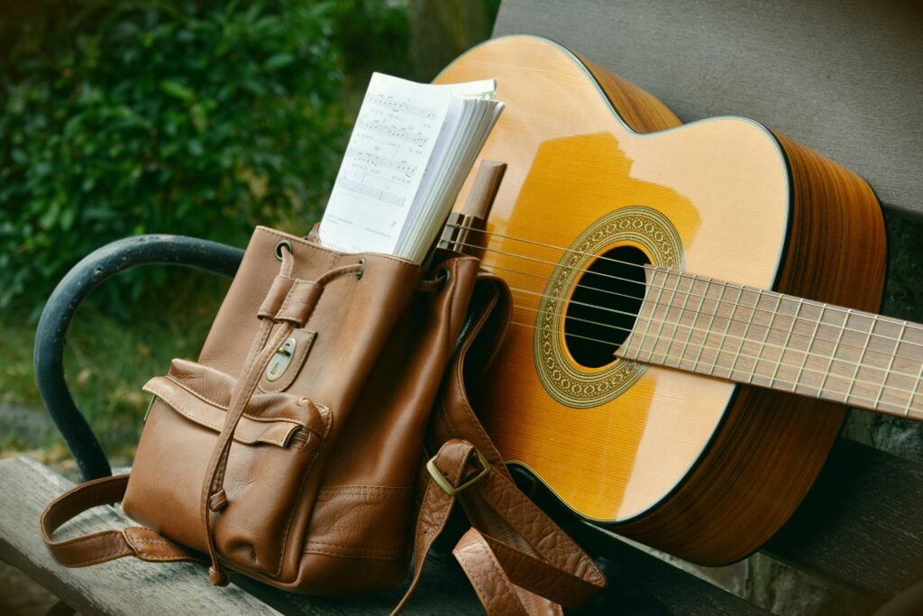 A classical guitar rests on a bench beside a brown leather backpack filled with sheet music and a wooden flute.