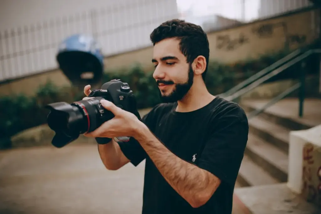 A man with a beard is standing outdoors, holding and examining a DSLR camera while wearing a black t-shirt. Steps and a fence are visible in the background.