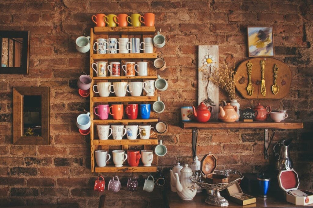 A rustic kitchen with a brick wall, shelves holding various colorful mugs, teapots, a metal colander, a serving tray, and assorted decorative items.