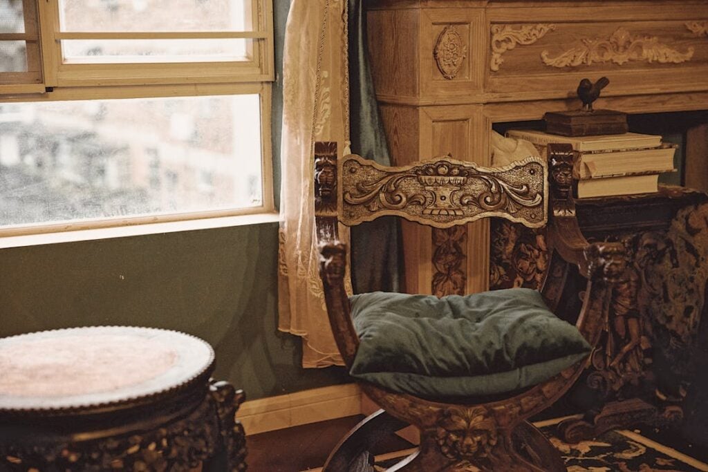 An antique wooden chair with ornate carvings and a blue cushion is placed beside a window. A round wooden table and an intricate wooden cabinet are also visible in the room.