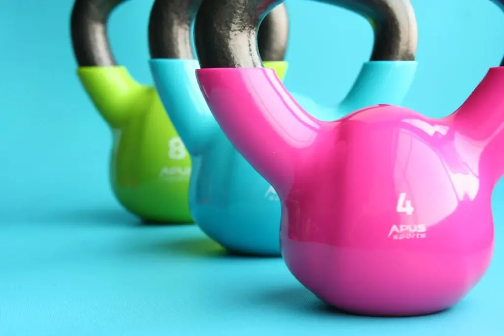 Three colorful kettlebells arranged in a row on a blue surface. The kettlebells are green (8 kg), blue (6 kg), and pink (4 kg).