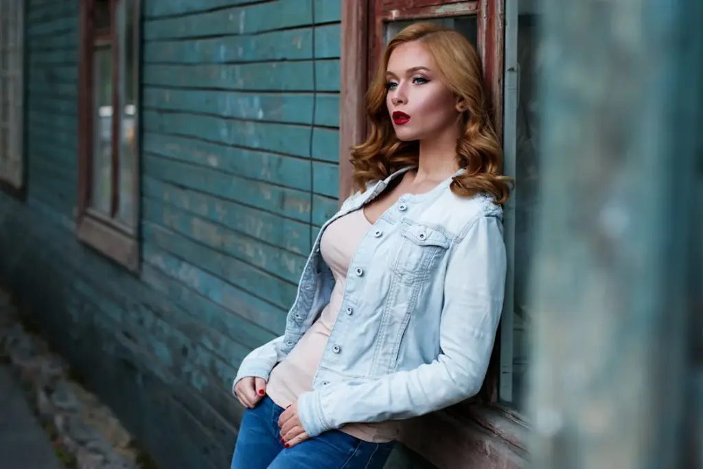 A woman with long hair and a light denim jacket leans against a wooden, blue-painted building. She gazes off into the distance.