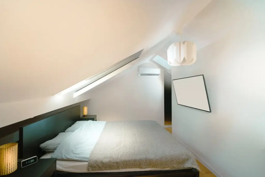 A minimalist bedroom with a sloped ceiling features a bed with a gray blanket, a wall-mounted TV, an air conditioner, a bedside lamp, and a digital clock showing 12:38.