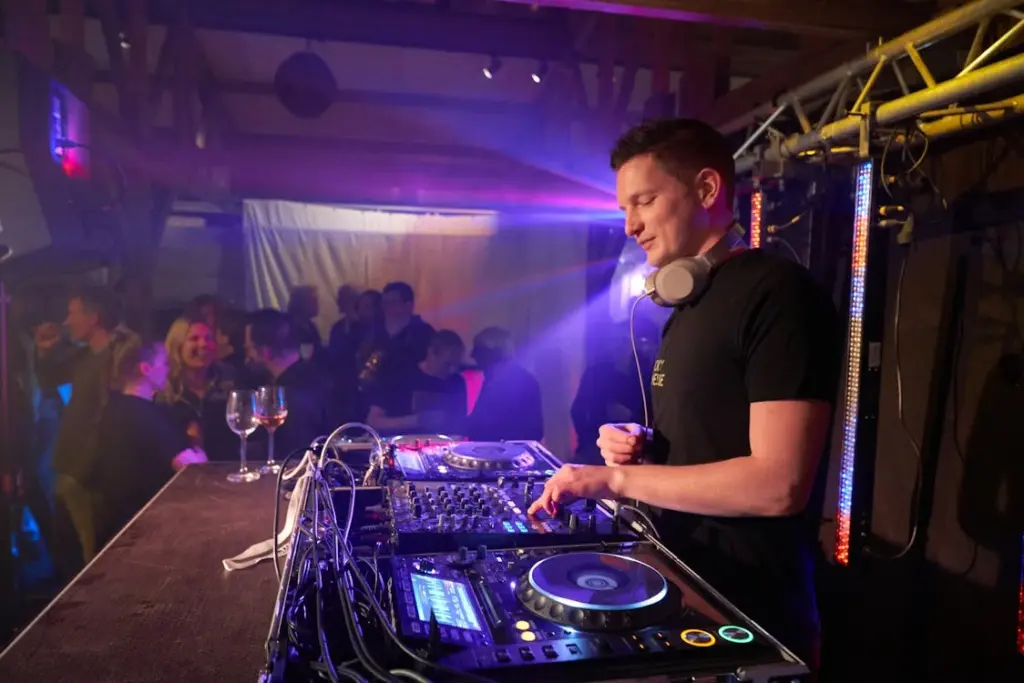 DJ performing at a nightclub, adjusting equipment on the console, with a crowd of people dancing in the background.