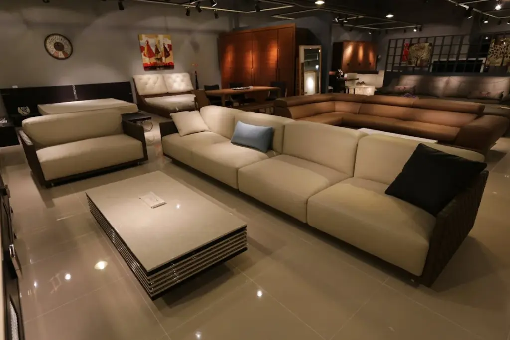 A showroom displaying various furniture pieces including a large beige sectional sofa, a white armchair, a gray coffee table, other sofas, and decor items in a modern, dimly lit setting.