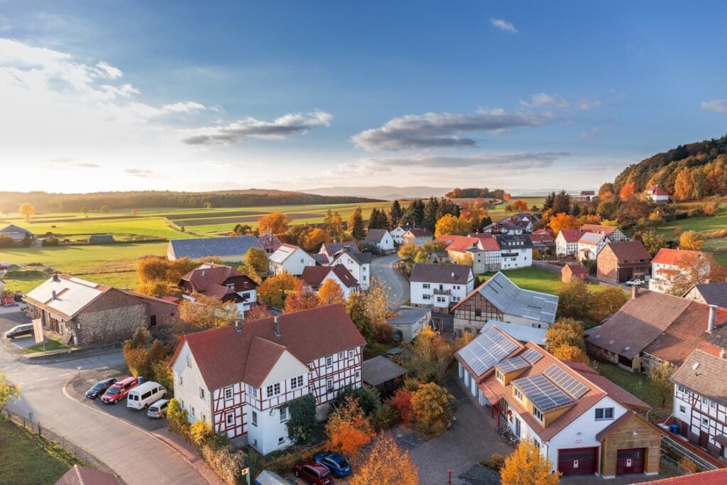 Aerial view of a small village with red-roofed houses surrounded by autumnal trees and green fields under a clear sky. Solar panels are visible on one building.