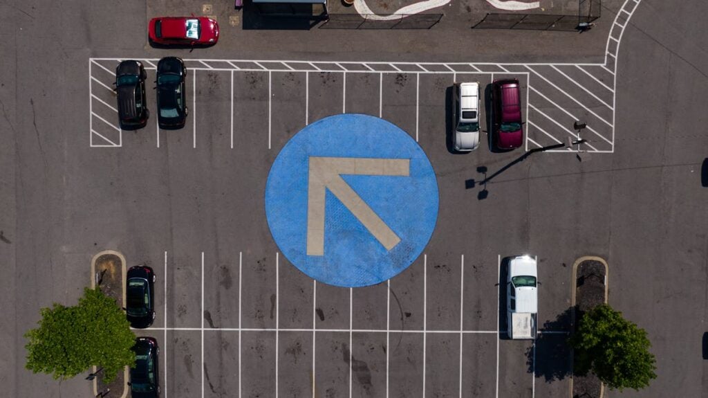 Aerial view of a parking lot with a large blue circular arrow pointing left in the center, several parked cars, and designated parking spaces visible.