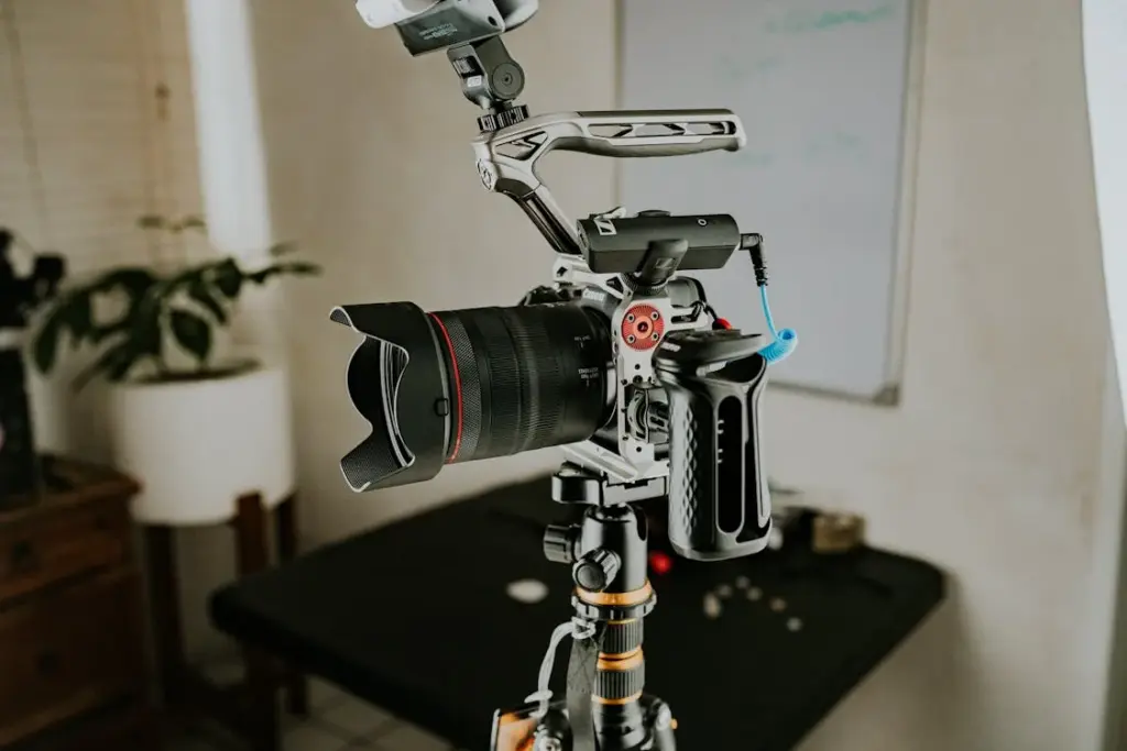 A professional camera mounted on a tripod stands in a room with a whiteboard and potted plant in the background.