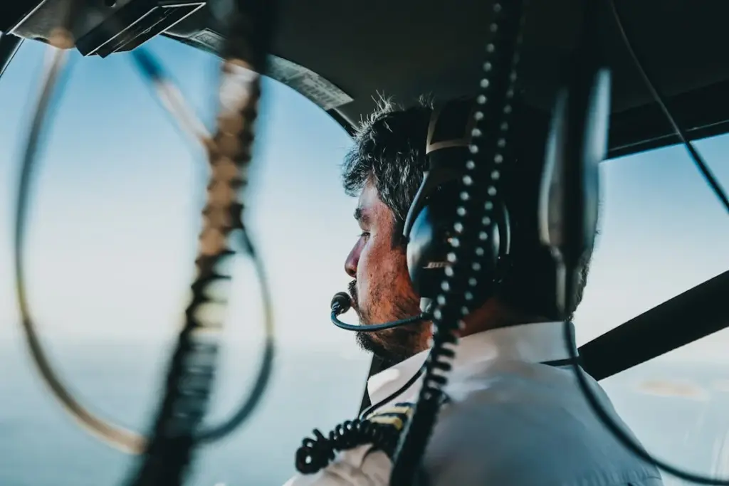 A pilot with a headset and microphone is seated in an aircraft cockpit, looking out of the window. Various cockpit instruments and controls are visible in the foreground.