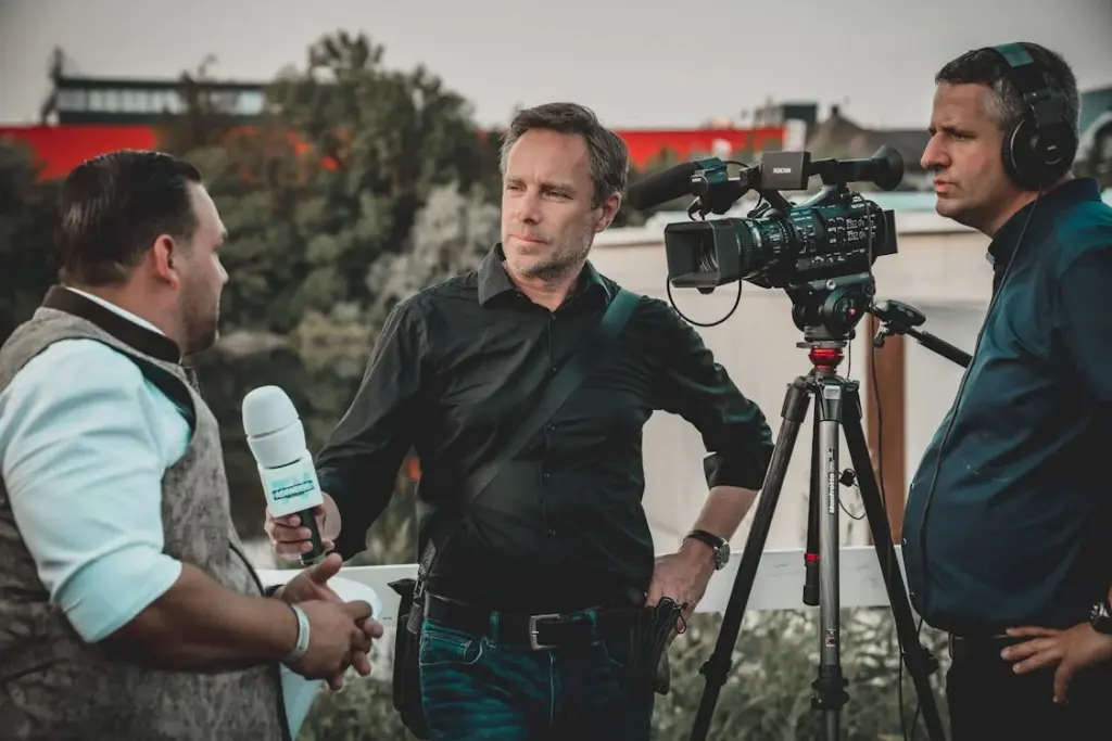 A journalist interviews a man while a cameraman records the conversation. Another individual wearing headphones stands near the camera. All are outdoors with trees in the background.