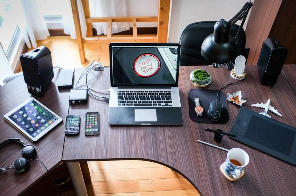 A cluttered desk with various gadgets including a laptop, tablet, smartphones, headphones, a camera, a smartwatch, and office supplies. A red button on the laptop screen reads 