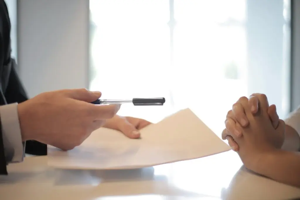 One person hands a document and a pen to another person whose hands are clasped together on a table.