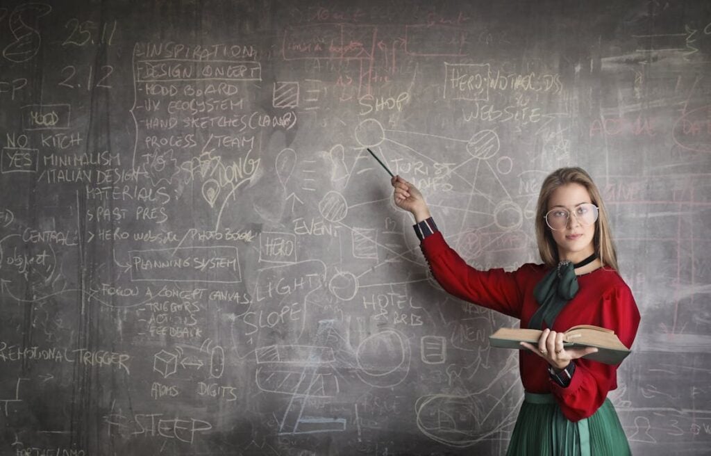 A woman in glasses and a red blouse stands in front of a chalkboard covered in drawings and notes, holding an open book in one hand and pointing at the chalkboard with a stick.