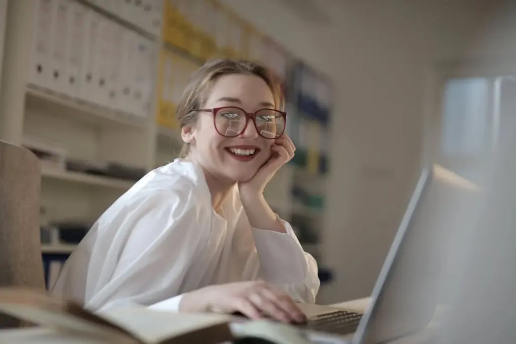 A person wearing glasses and a white shirt is sitting at a desk, smiling, and looking at a laptop. Shelves with files and books are in the background.