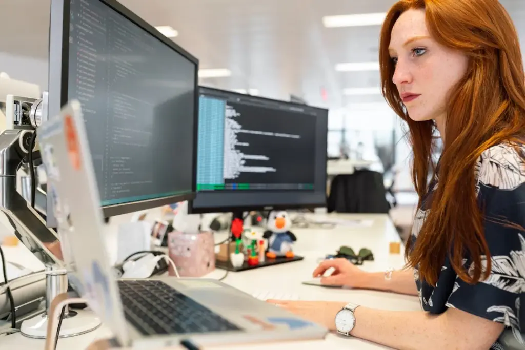 A person with long red hair working at a desk with multiple computer monitors displaying code in an office setting.